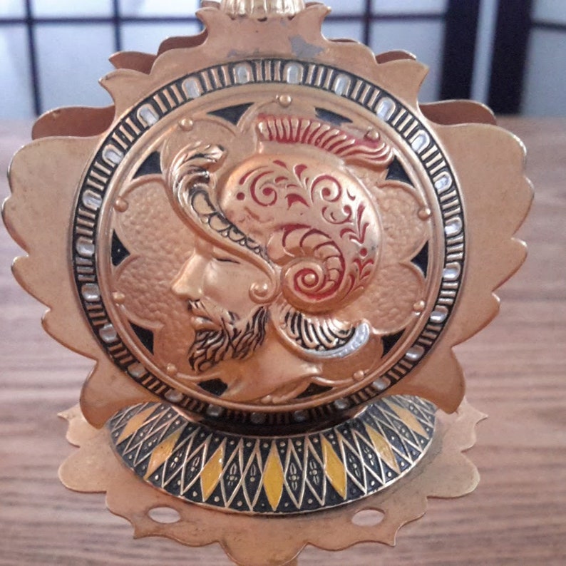 May include: Gold metal napkin holder with a decorative design of a warrior's head in a helmet. The design is in black, red, and gold.