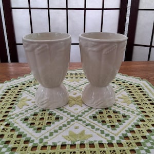 Peut inclure: Deux vases en céramique blanche avec un motif floral en relief. Les vases ont une base large et un col étroit. Ils sont posés sur un napperon en crochet vert et blanc.