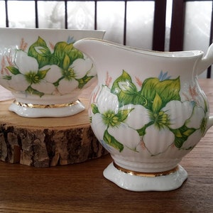 Peut inclure: Un ensemble de crémier et de sucrier en porcelaine blanche avec un motif floral composé de fleurs blanches et de feuilles vertes. L'ensemble est orné d'une bordure dorée.