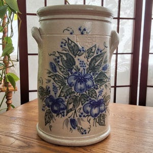 May include: A cylindrical beige stoneware crock featuring two side handles and a hand-painted floral design in shades of blue and green. The front displays a stamped logo reading Salmon Falls Stoneware, with blue painted flowers and leaves.