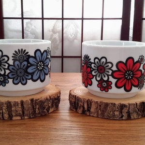 May include: Two white ceramic mugs with floral designs. The mug on the left has blue flowers and the mug on the right has red flowers. Both mugs are sitting on wooden coasters.