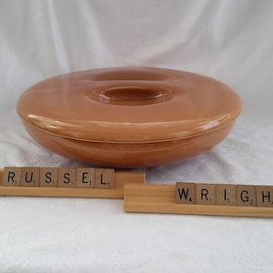 Peut inclure: Un plat à casserole rond en céramique brun clair avec un couvercle. Le plat a une poignée centrale sur le couvercle. Le nom "RUSSELL WRIGHT" est épelé en tuiles Scrabble en dessous.