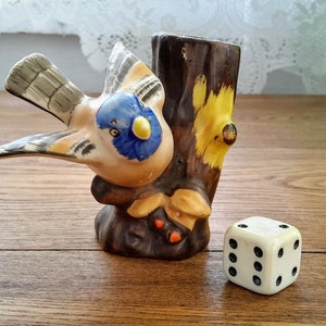 Peut inclure: Une figurine en céramique d'un oiseau bleu perché sur un tronc d'arbre. L'oiseau a un bec jaune et des marques bleues et brunes. Le tronc d'arbre est peint en brun et jaune. Un dé blanc avec des points noirs est au premier plan.