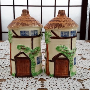 May include: A pair of ceramic salt and pepper shakers shaped like cottages with thatched roofs. The cottages are painted in shades of brown, white and blue with green ivy.