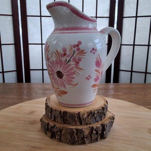 May include: A white ceramic jug with a pink rim and floral design. The jug is sitting on a wooden coaster.