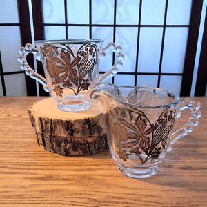 May include: A clear glass sugar bowl with two beaded handles and a matching creamer pitcher, both decorated with metallic gold floral patterns. The sugar bowl features two handles, while the creamer has a single handle and a pouring spout.