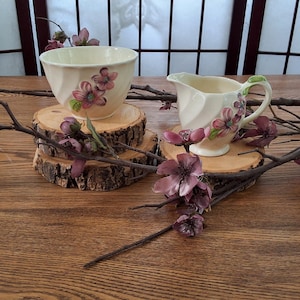May include: Cream-coloured teacup and creamer set with pink floral accents. The teacup and creamer are displayed on wooden coasters, with decorative branches and flowers. The set is on a wooden table.