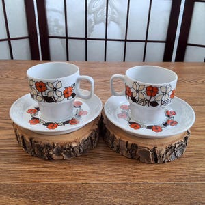 May include: Two white ceramic teacups and saucers with a floral design in orange, black, and white. The cups and saucers are placed on small, round wooden coasters. The background includes a wooden table and a window.