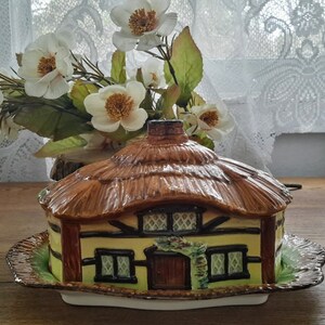 May include: A ceramic cottage-shaped butter dish with a thatched roof, brown trim, and green accents. The dish is on a white and green plate.