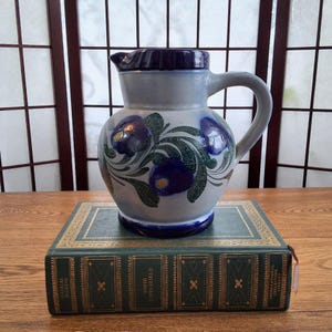 Peut inclure: Une cruche en céramique avec un bord et une anse bleus, ornée de motifs floraux bleus et de feuilles vertes. La cruche repose sur un livre vert avec des lettres dorées. L'arrière-plan présente un paravent en bois.