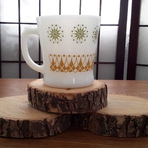 May include: A white ceramic mug with a green and gold geometric design sits on top of three wooden coasters. The coasters are made from tree bark and have a natural, rustic look.