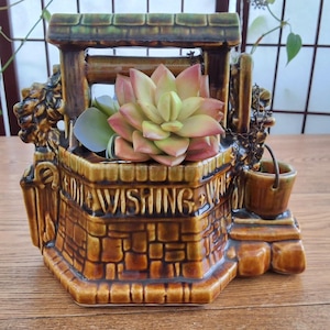 Peut inclure: Un pot de fleurs en céramique en forme de puits à souhaits, dans les tons de marron et de vert, avec une fausse plante succulente à l'intérieur. Le pot a une texture de brique et un petit seau suspendu à une chaîne. Les mots "For Wishing Well" sont inscrits sur le devant.
