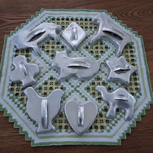 May include: A set of ten vintage metal cookie cutters in various shapes, including a star, heart, camel, chicken, horse, and more. The cutters are arranged on a white doily with a green and white checked pattern.