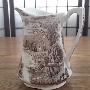 Peut inclure: Un pichet en céramique blanche avec un motif de paysage brun et blanc. Le pichet a une anse et un bec verseur. Le motif de paysage représente une maison, des arbres et une rivière.