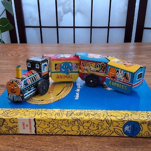 May include: Vintage toy train set with a colorful locomotive and three cars. The cars are labeled "ANIMAL" and "EXPRESS" and feature illustrations of animals. The train is made of metal and sits on a blue surface.