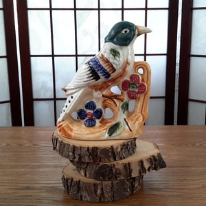 May include: A ceramic bird figurine with blue, green, white, and red painted details, perched on a branch with flowers. The figurine is displayed on a stack of three wooden slices.