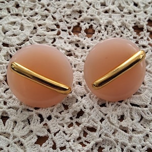 Peut inclure: Une paire de boucles d'oreilles circulaires dotées d'une base en résine rose pâle lisse. Chaque boucle d'oreille est rehaussée d'une barre en métal doré poli qui s'étend en diagonale sur le centre de la surface ronde, créant un design équilibré.