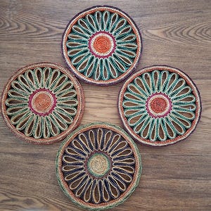 Peut inclure: Quatre dessous de verre circulaires tressés présentant un motif en spirale central entouré d'un anneau de boucles en forme de pétales. Les dessous de verre affichent une palette de couleurs vives orange, rouge foncé, vert forêt et beige.