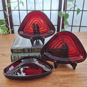 May include: Three triangular, ruby-red glass dishes. Each dish has a unique shape and is supported by a black stand. The dishes are arranged on a wooden surface, with antique books in the background. The glass has a layered design.