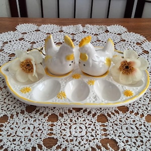 May include: A white ceramic egg holder with a yellow trim, featuring two chicken figurines and spaces for eggs. The holder is decorated with yellow floral accents and is accompanied by white flowers.