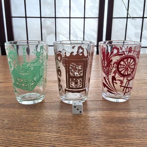 May include: Three clear glass tumblers with different vintage-inspired designs. The tumblers feature a green, brown, and red design. The green tumbler has a design of a sewing machine, the brown tumbler has a design of a clock, and the red tumbler has a design of a spinning wheel.