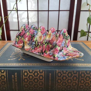 May include: A ceramic toast rack with a floral pattern. The rack has six slots and is decorated with pink, red, and blue flowers on a black background. The base is white with a gold trim. It is placed on a vintage book.
