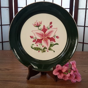 May include: A white ceramic dinner plate with a green rim and a gold trim. The plate features a pink and red flower with green leaves.