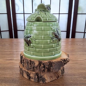 May include: A light green ceramic honey pot shaped like a traditional woven beehive. The exterior features a textured basket-weave pattern with two sculpted bees attached to the sides and a lid with a small bee handle and a notch for a spoon.
