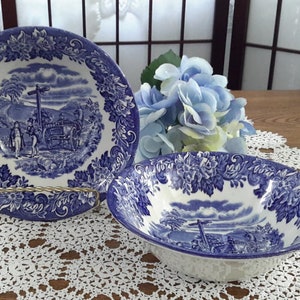 Peut inclure: Deux bols en céramique bleu et blanc avec un motif floral et une scène de calèche sur une route de campagne. Les bols sont sur une dentelle blanche au crochet.