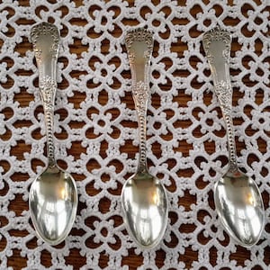 Peut inclure: Quatre cuillères à café en argent avec des manches ornés et des bols polis. Les cuillères sont disposées en rangée sur un napperon en crochet blanc.