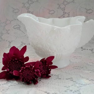 Peut inclure: Une crémier en verre de lait blanc avec un bord festonné et un motif floral. La crémier est posée sur une nappe en dentelle blanche avec trois fleurs bordeaux.