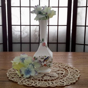May include: A white porcelain vase with a floral and bird design. The vase is decorated with pink, blue, and green flowers and a blue bird. The vase is sitting on a white lace doily on a wooden table.