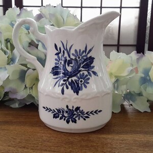 Peut inclure: Un pot à crème en céramique blanche avec un motif floral bleu. Le pot à crème a une poignée et un bec verseur. Il est posé sur une table en bois.