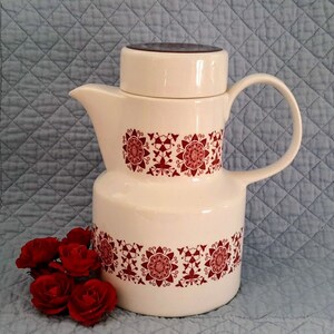 Peut inclure: Une cafetière en céramique blanche avec un motif géométrique rouge et blanc. La cafetière a un couvercle et une poignée. La cafetière est posée sur une surface matelassée bleue.