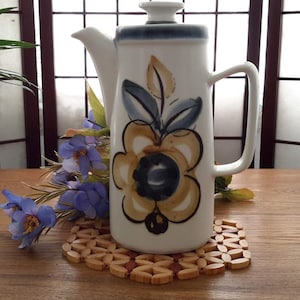 Peut inclure: Une cafetière en céramique blanche avec un motif floral bleu et brun. La cafetière a un couvercle et une poignée. Elle est posée sur un dessous-de-plat en bois.