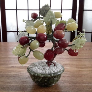 Peut inclure: Un arbre à raisin en jade avec des raisins rouges et verts, dans un pot vert avec des pierres blanches. L'arbre est sur une surface en bois.