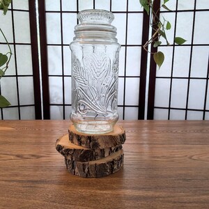 Peut inclure: Un bocal en verre transparent avec un couvercle, orné d'un motif de blé en relief. Le bocal est présenté sur une pile de trois dessous de verre en bois. L'arrière-plan comprend un écran en bois et de la verdure.