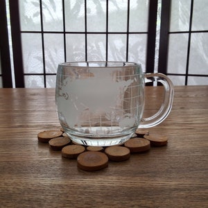 Peut inclure: Une tasse en verre transparent avec une carte du monde gravée sur le côté, posée sur un dessous-de-verre en bois fait de rondins de bois empilés.
