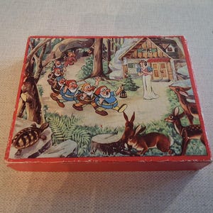May include: A vintage wooden box with a red frame and a colourful illustration of Snow White and the Seven Dwarfs. The scene depicts the dwarfs walking through a forest towards a cottage, with Snow White and forest animals.