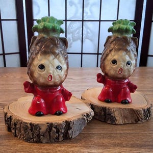 May include: Two ceramic pineapple-shaped figurines with human-like features. They have brown and tan bodies, green leafy tops, and wear red outfits. Each figurine sits on a wooden slice.