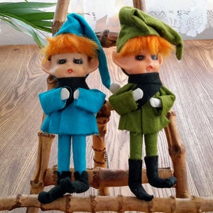 Peut inclure: Deux poupées d'elfes vintage aux cheveux roux, l'une vêtue d'une tenue bleue et l'autre d'une tenue verte. Les deux poupées ont des bottes noires et sont assises sur un support en bois.