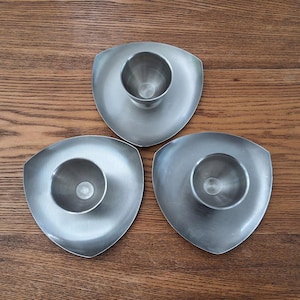 May include: Three brushed metal egg cups with a triangular base. Each cup has a rounded center for holding an egg. The cups are arranged on a wooden surface.