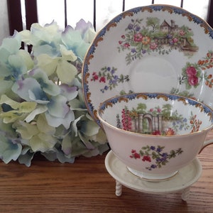 May include: A vintage teacup and saucer set with a floral and garden scene design. The cup and saucer are white with gold trim and feature a blue and gold border. The floral design includes pink, red, and yellow flowers.