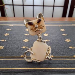 Peut inclure: Broche vintage en forme de chat. La tête et le corps du chat sont carrés et de couleur beige clair. Il présente des yeux rouge foncé et des moustaches dorées. La broche est en métal doré et a une queue incurvée.