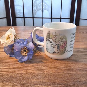 Peut inclure: Un mug en céramique blanche orné d'une illustration colorée de lapins et du texte Once upon a time there were four little Rabbits and their names were Flopsy Mopsy Cotton-tail and Peter. À côté du mug se trouvent des fleurs artificielles blanches et violettes.