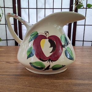 Peut inclure: Pichet en céramique crème avec un grand motif de pomme rouge. La pomme a un centre jaune et est entourée de feuilles vertes. La poignée et le bec sont également crème, avec des touches de rouge et de bleu sur la poignée.