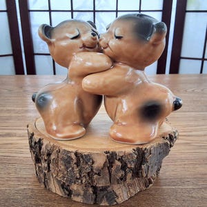 May include: Two glossy ceramic bear figurines in a light brown finish with dark brown accents on the ears and backs. The bears are positioned facing each other in an embrace, with closed eyes and smiling expressions, standing on a round wood slice.