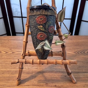 Peut inclure: Un vase mural gris foncé texturé orné de motifs floraux en relief rouge orangé et de feuilles vertes, présenté sur un support trépied en bambou brun clair. Une plante retombante aux feuilles vertes et blanches panachées sort du vase.