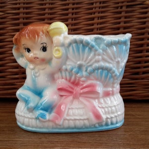 May include: A ceramic planter shaped like a baby in a blue and white basket with a pink bow. The baby is holding a yellow rattle.