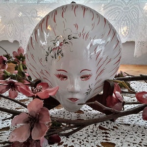 May include: A white ceramic wall mask with red and black painted details. The mask features a stylised female face with closed eyes and a delicate floral design. The mask is displayed on a wooden stand.